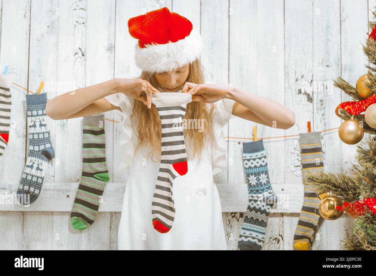 Christmas Stockings. Curious Girl in a White Dress and a Santa Hat on her Head Looks into an Empty Striped Sock. Close-up Stock Photo
