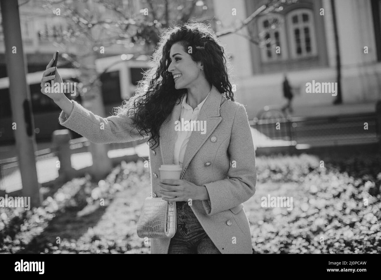 Pretty young woman using smartphone to take selfie photo on the street and holding takeaway coffee Stock Photo