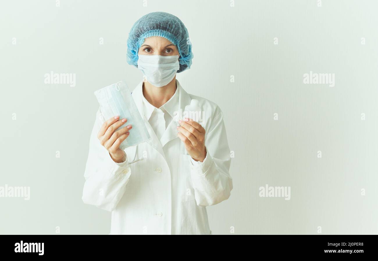 Female doctor in medical mask and uniform offers mask and disinfectant in bottle Stock Photo