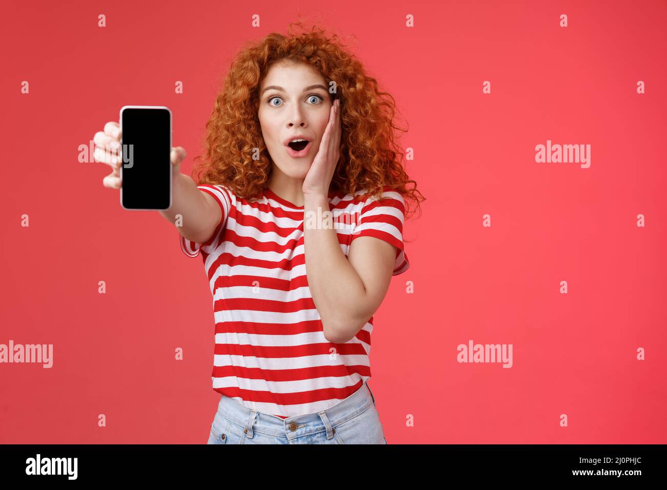 Amused impressed redhead good-looking curly-haired excited girl share awesome social media page show smartphone screen amazed to Stock Photo