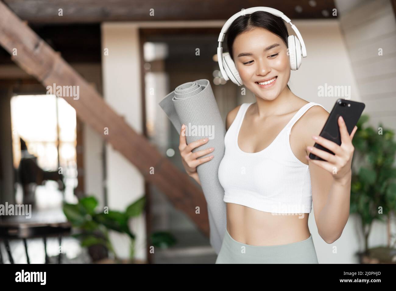 Smiling asian girl with headphones, looking at mobile phone and holding rubber mat for workout, fitness at home, concept of life Stock Photo