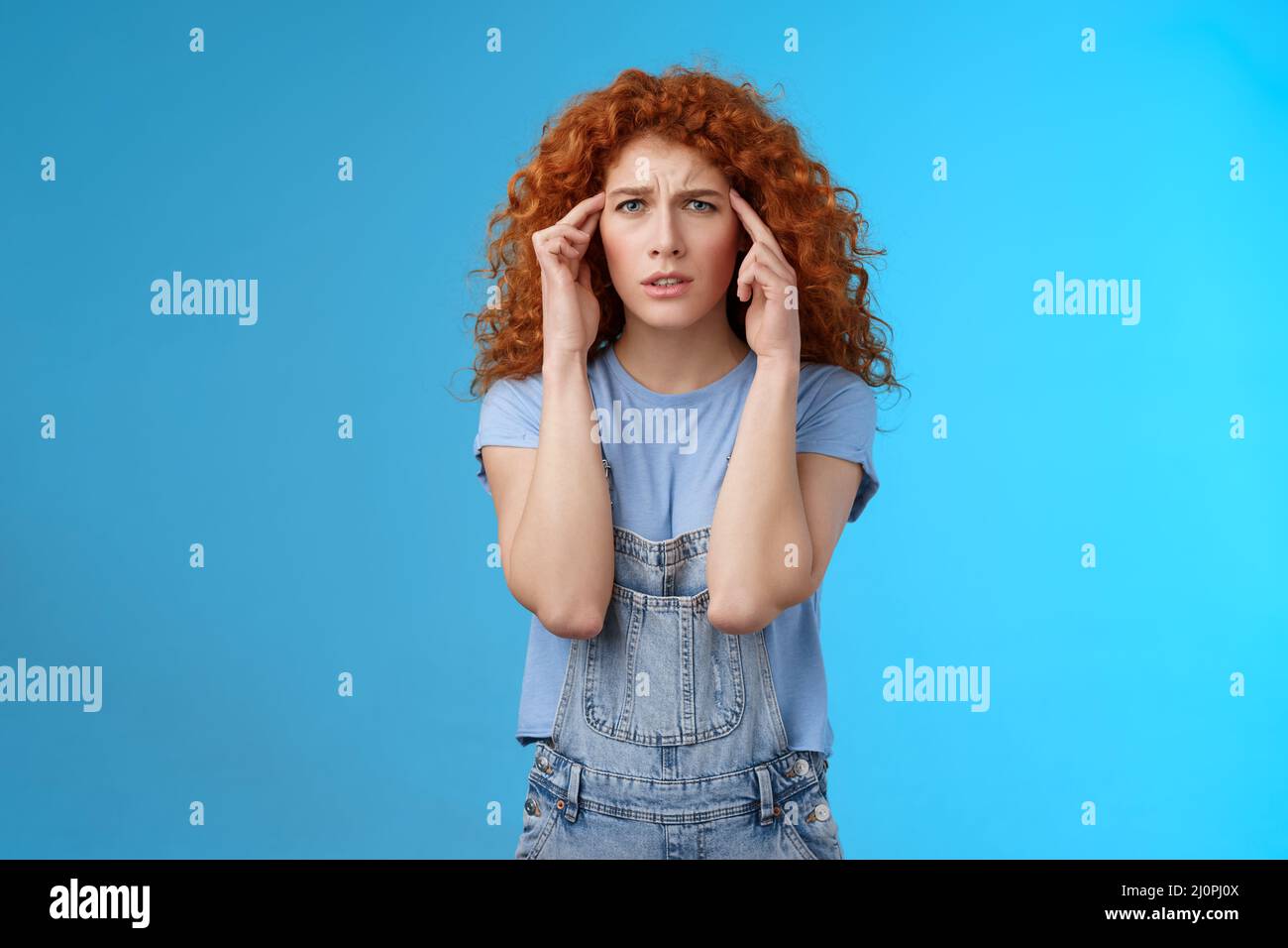Confused panicking young distressed redhead curly girlfriend frowning touch temples cannot focus look perplexed worried not reme Stock Photo