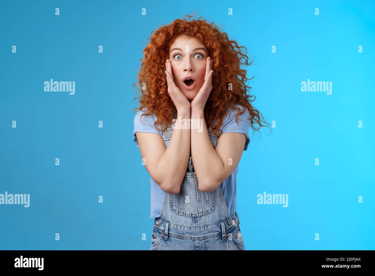 Amazed gasping shocked wondered redhead attractive curly girl popping eyes stare camera drop jaw concerned astonished touch chee Stock Photo