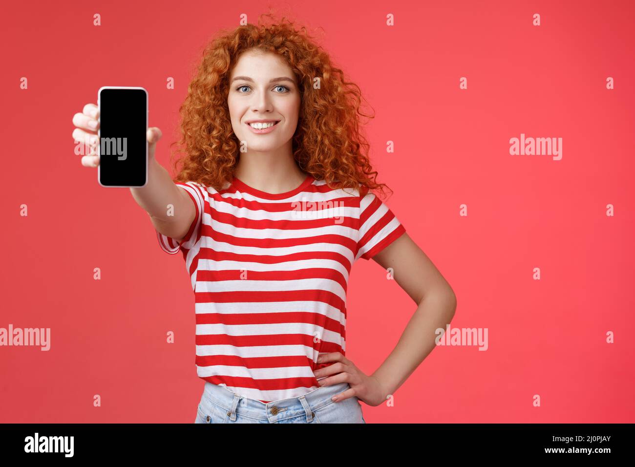 Confident good-looking redhead curly woman present cool app hold smartphone extended hand show phone screen smiling assertive re Stock Photo
