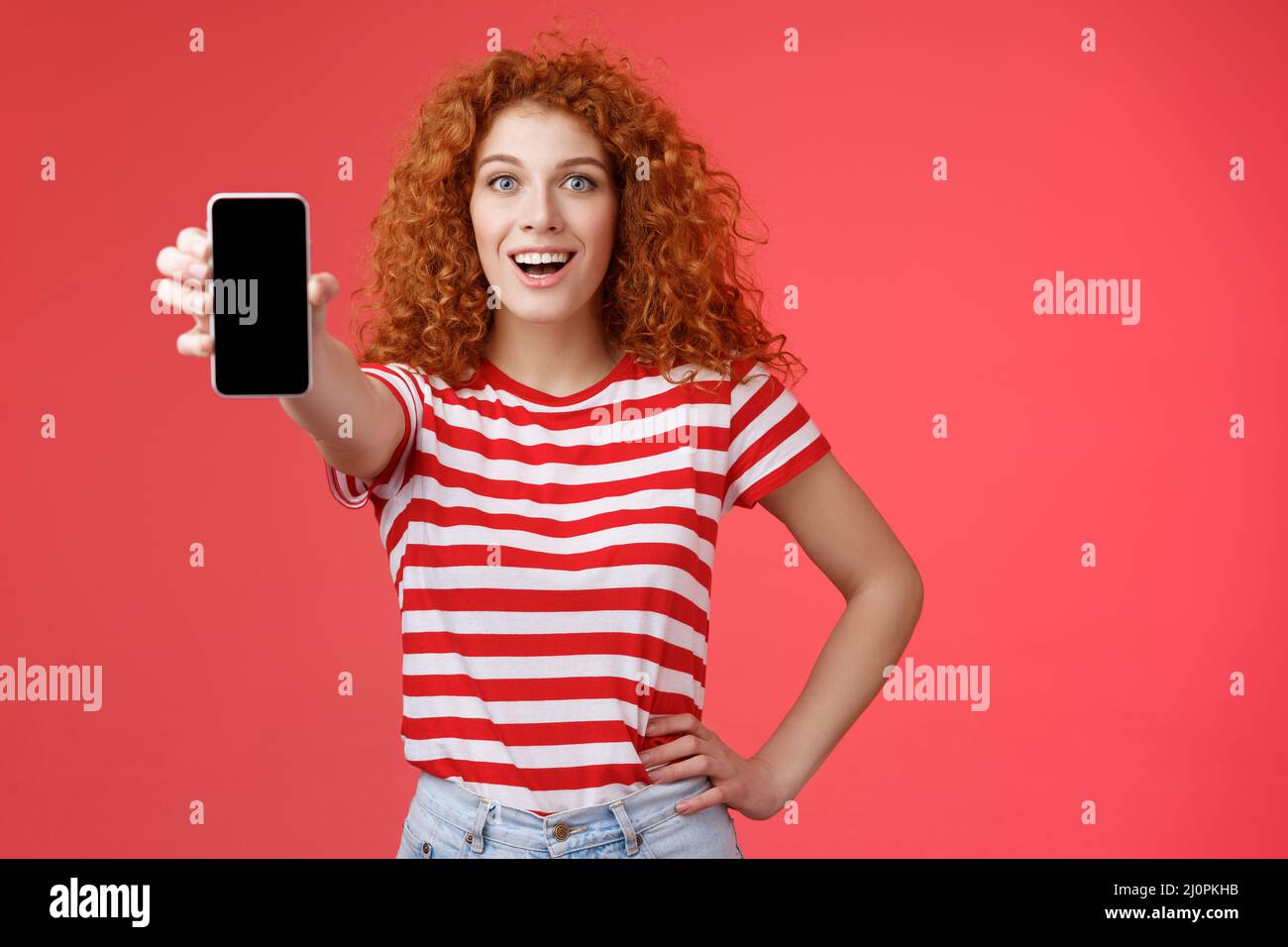 Cheerful attractive charismatic european redhead girl curly hairstyle show smartphone screen smiling happily promote app advice Stock Photo