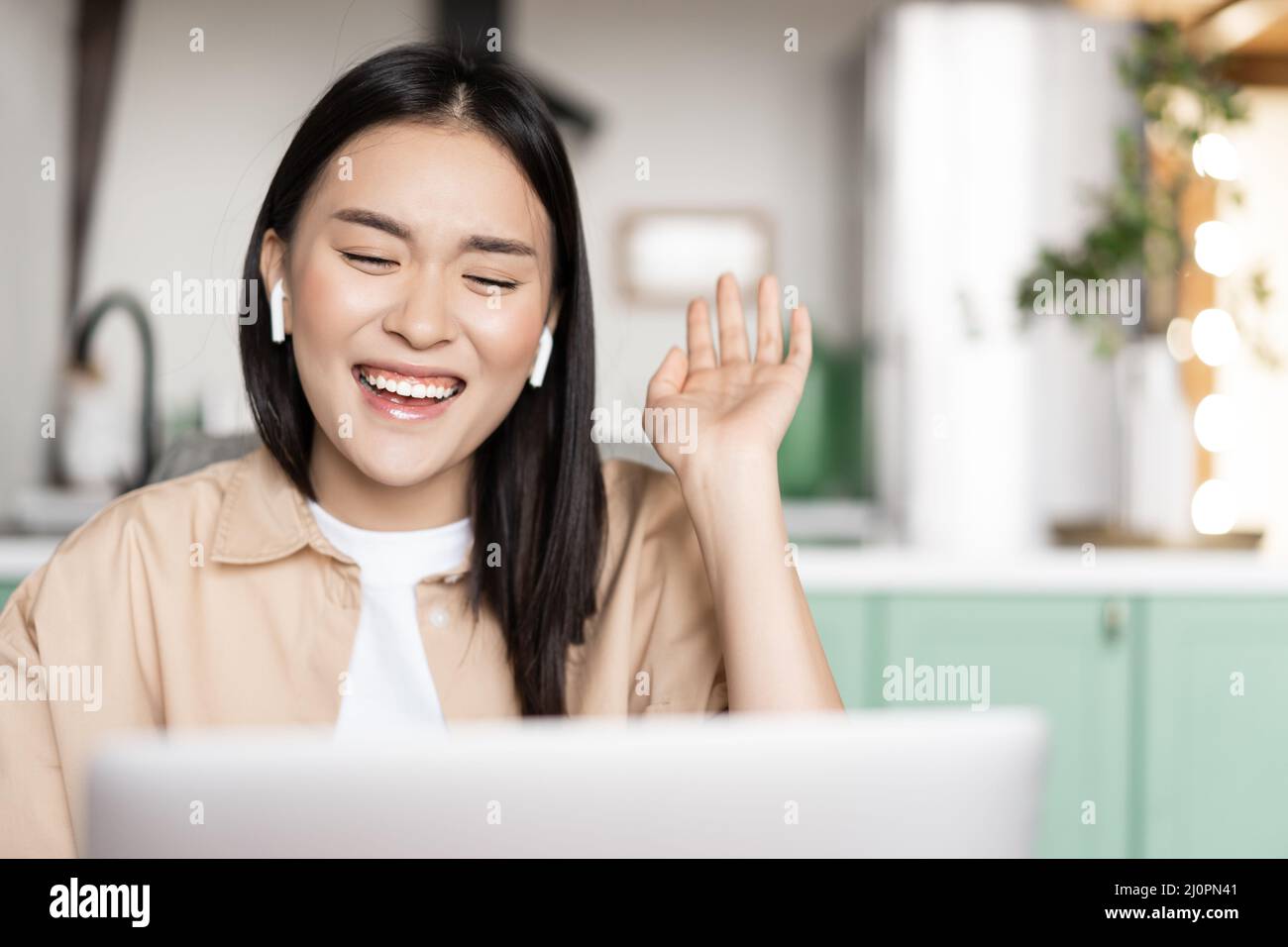 Happy asian woman laughing and smiling, talking on video call using laptop, waving hand and chuckling, wearing earbuds Stock Photo