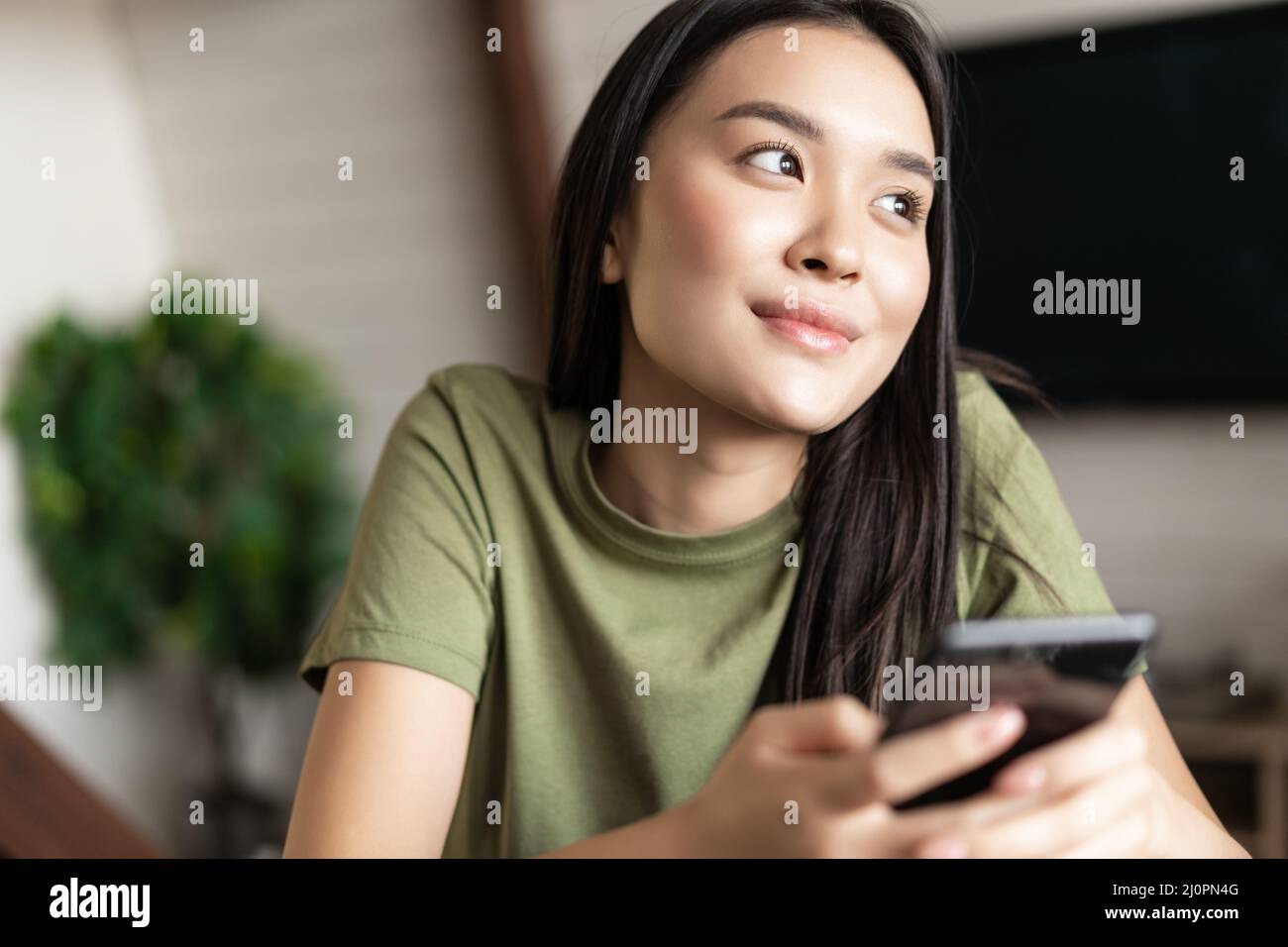 Young dreamy woman looking outside window, holding smartphone and smiling, chatting on phone app Stock Photo