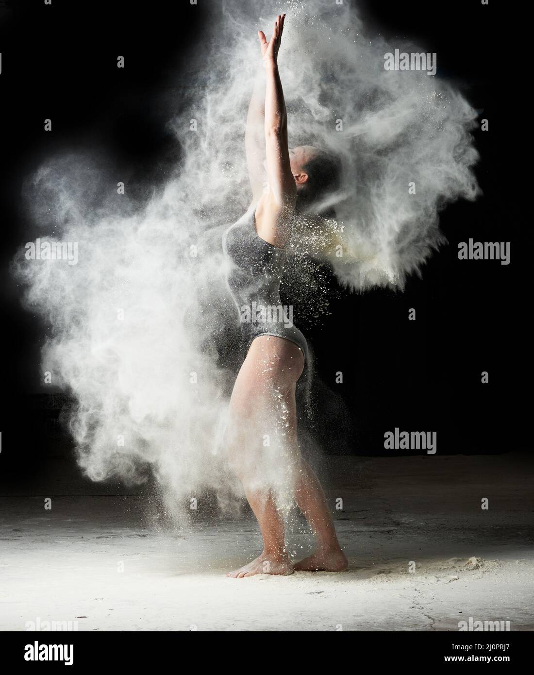 Beautiful caucasian woman in a black bodysuit with a sports figure is dancing in a white cloud of flour on a black background Stock Photo