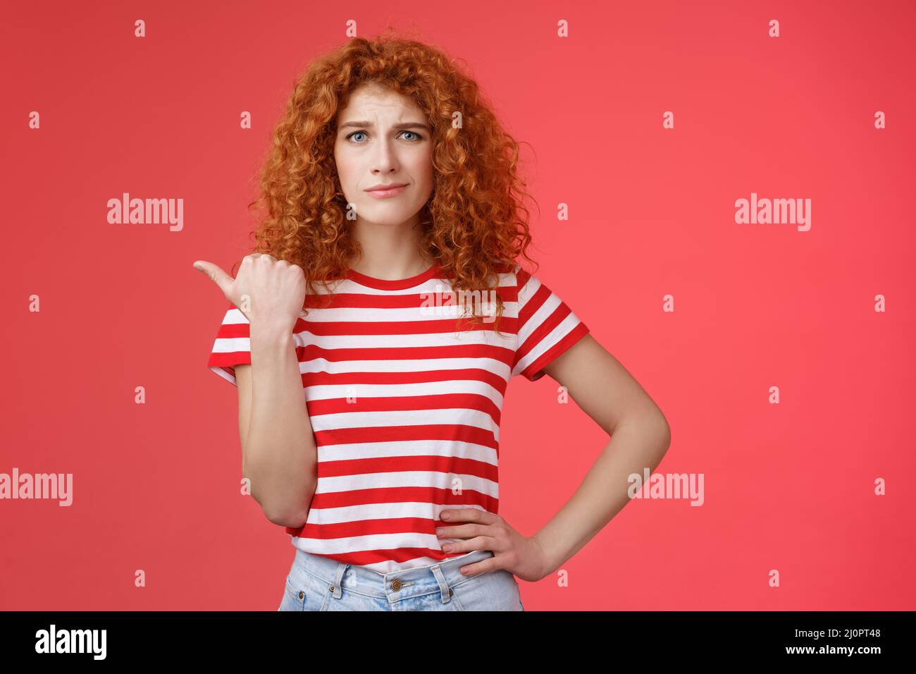 Girl not sure best choice. Doubtful redhead curly woman look suspicious frowning uncertain stare hesitant pointing left thumb un Stock Photo