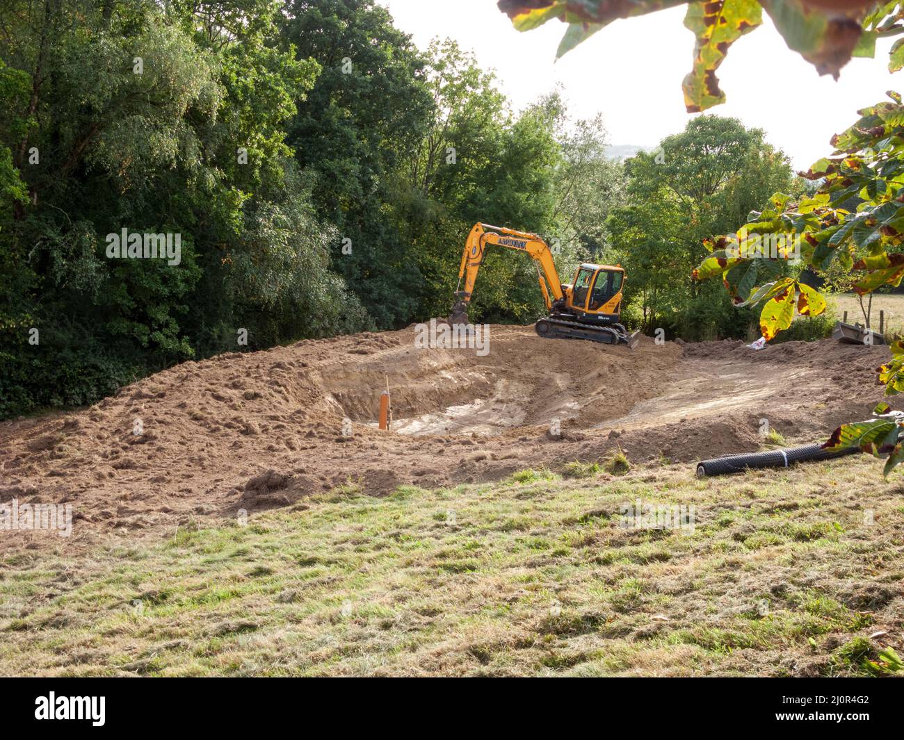 UK, England. Pond construction on a sloping field. 10 x 5 meters. Stage four - digging down to depth of 1.5 meters & creating a bund. Overflow pipe install Stock Photo