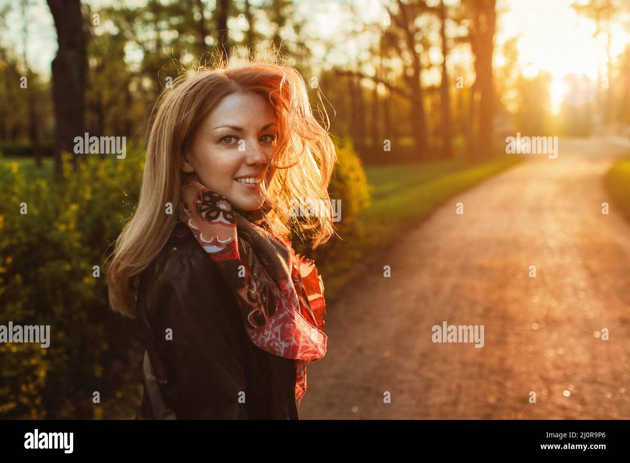 Young smiling woman with waving hair on park road at sunset. Girl walking in evening forest. Stock Photo