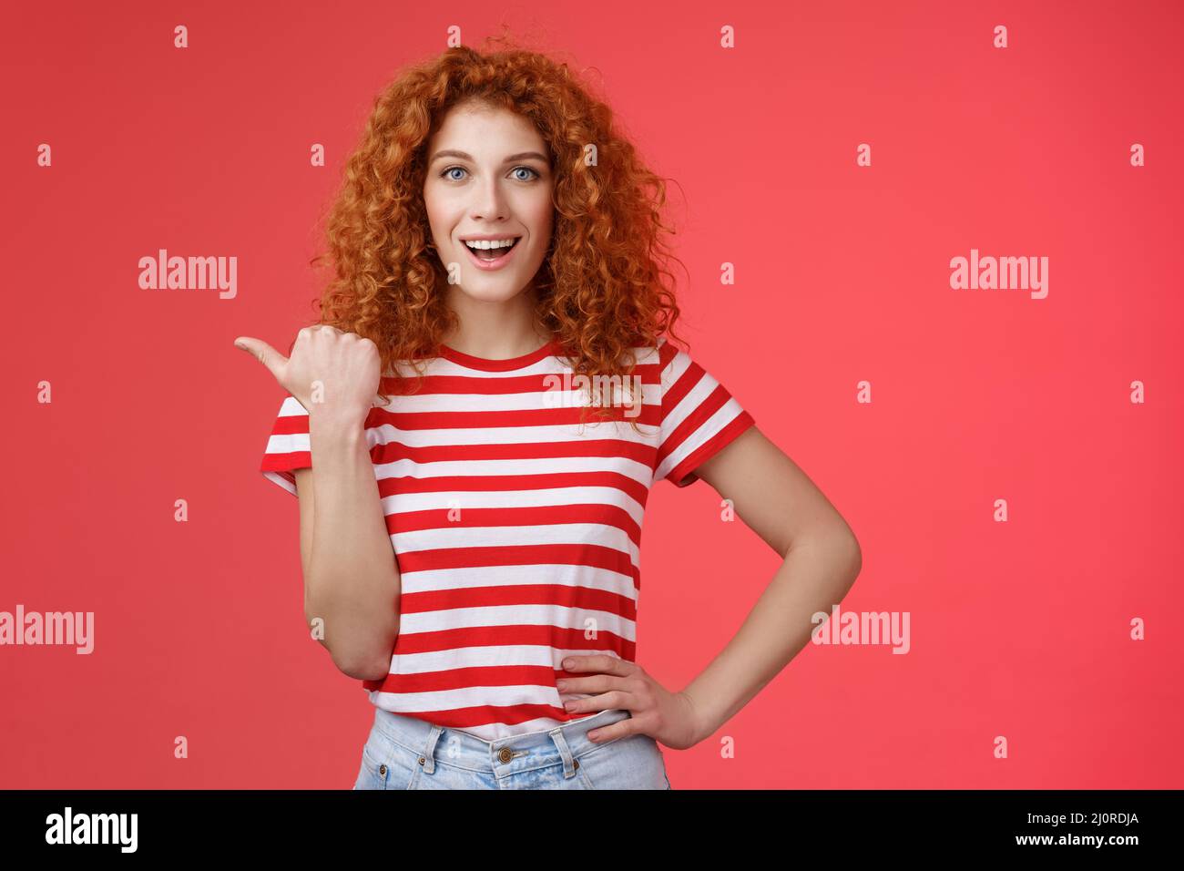 Sassy good-looking emotive happy smiling redhead european female curly hairstyle pointing thumb left grin assertive cheeky hold Stock Photo