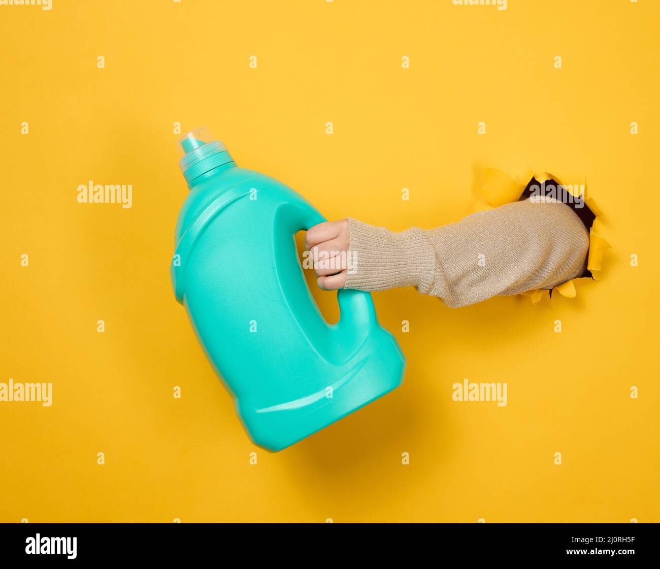 Blue plastic bottle with liquid detergent in a female hand on a yellow background. A part of the body sticks out of a torn hole Stock Photo
