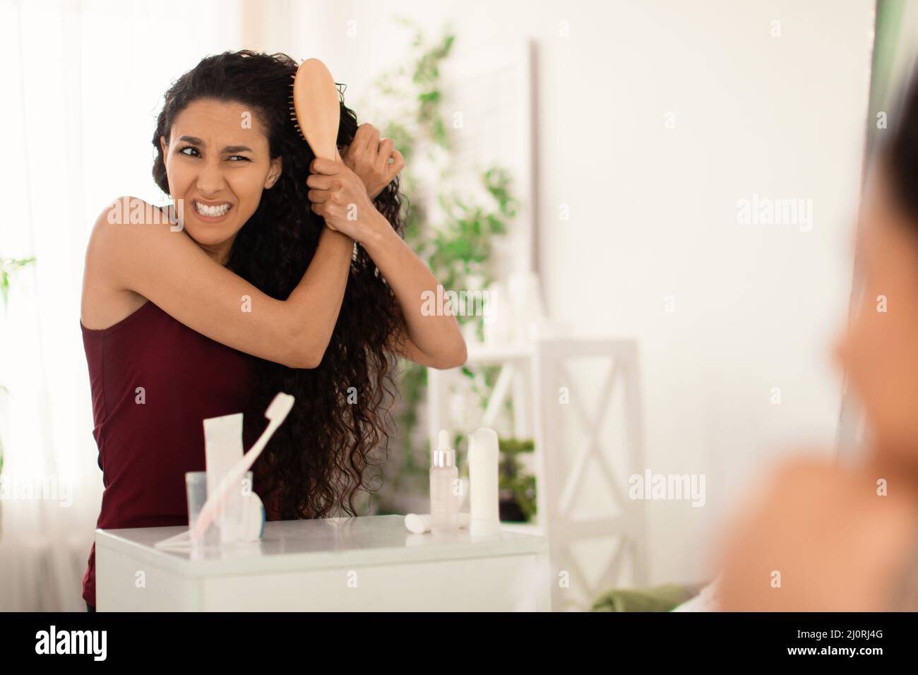 Frustrated millennial female having problem brushing her tangled hair near mirror at bathroom, blank space Stock Photo