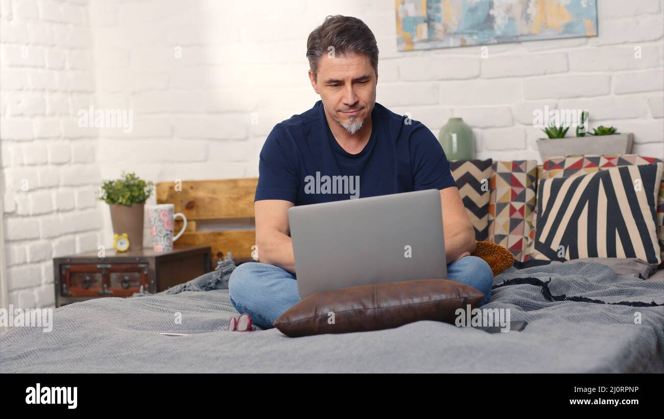 Casual man sitting on bed working online from home with laptop computer