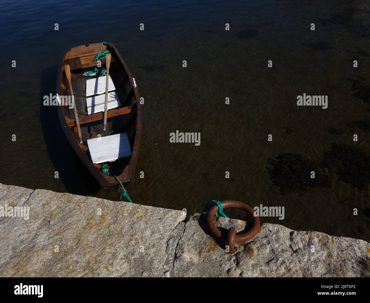 A small rowing boat with the oars lying on the thwart moored to an old stone quay, a stone breakwater, in a small fishing harbour and village. Stock Photo