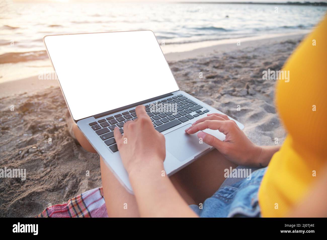 Close-up of a cut-out laptop screen on the lap of a young caucasian woman sitting on a sandy beach by the sea at sunset. Freelan Stock Photo