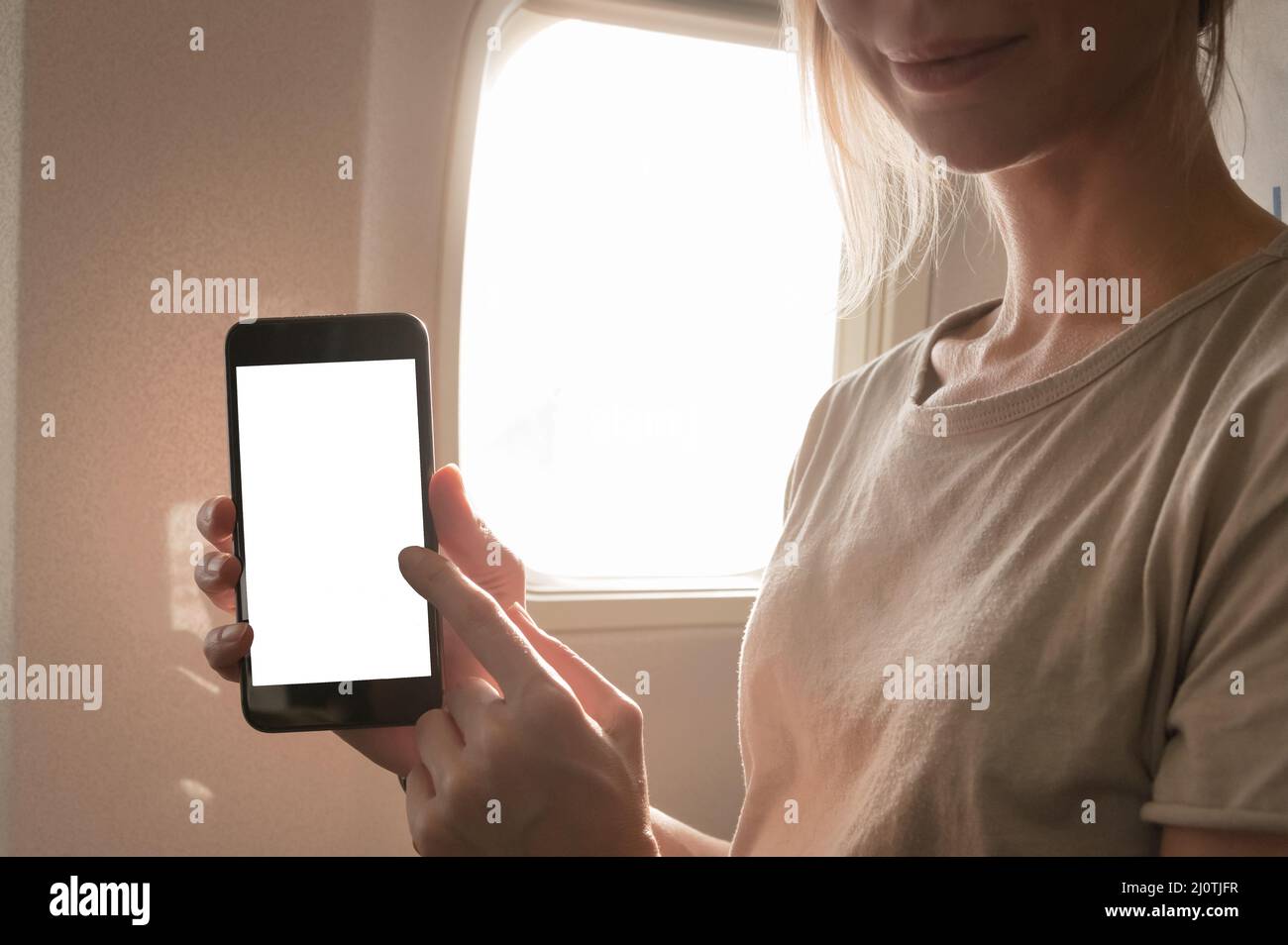 Mockup image of a woman holding and looking at smart phone with blank white screen next to an airplane window with clouds and sk Stock Photo