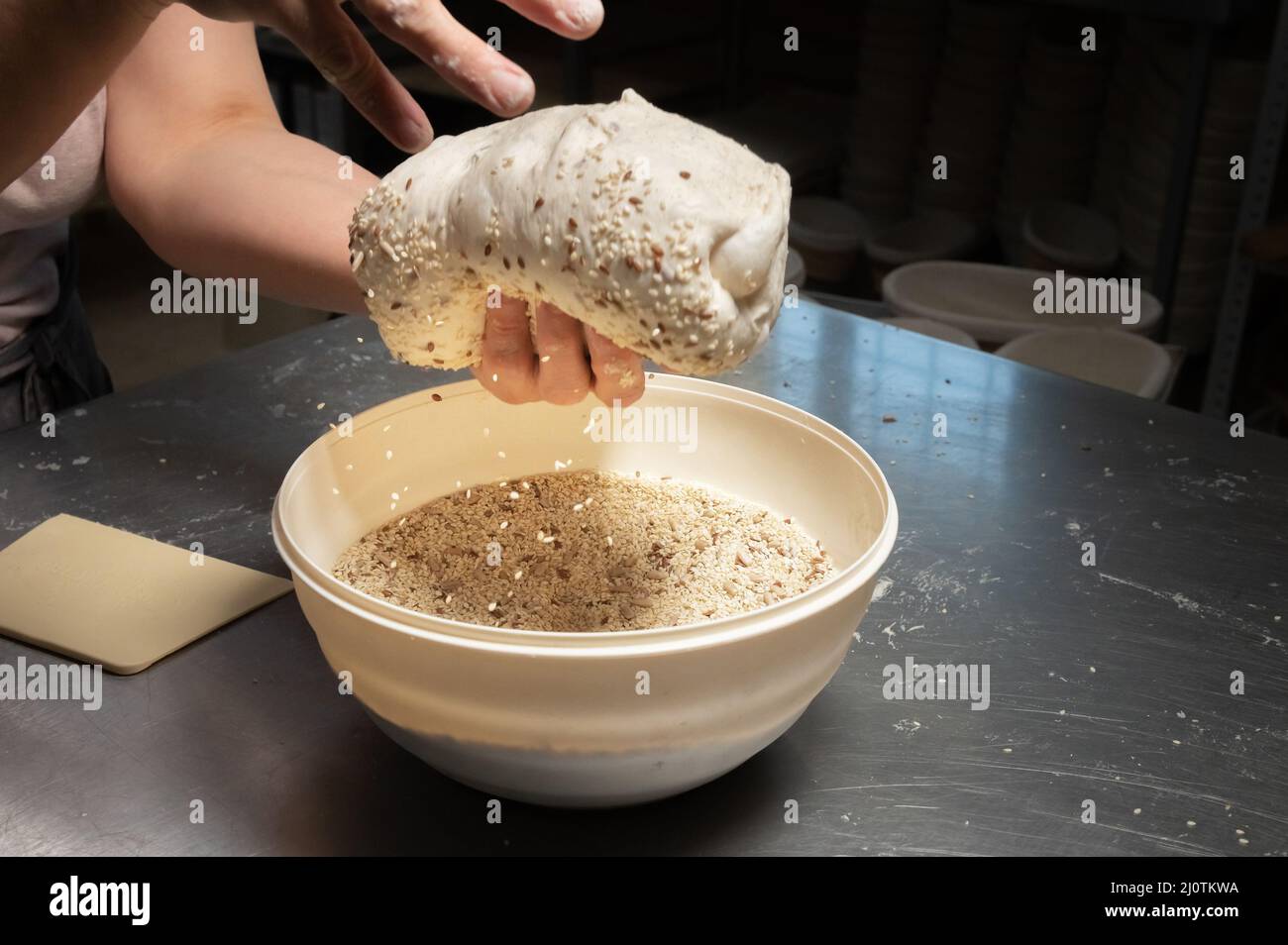 Close-up of a baker's female hands dipping dough into a bowl of seeds and cereals before baking artisan bread at a home bakery Stock Photo