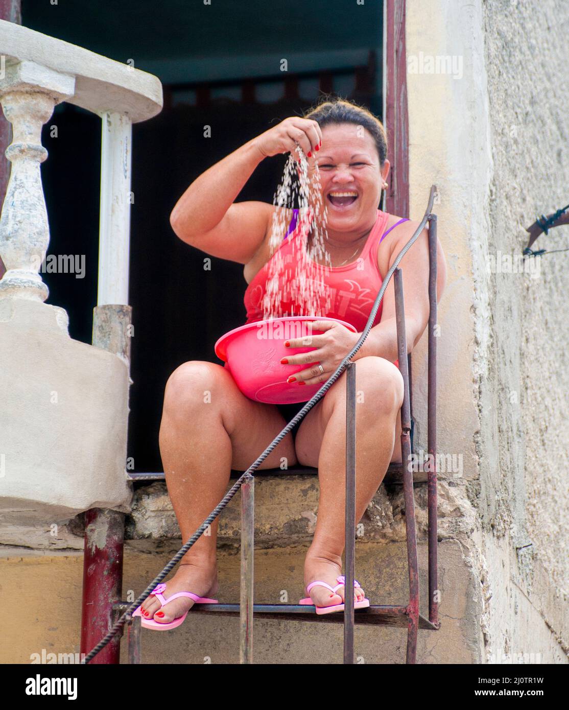 Woman sitting on a rot iron staircase in front of her home in Camagüey, Cuba preparing white rice in a bowl for dinner. Stock Photo