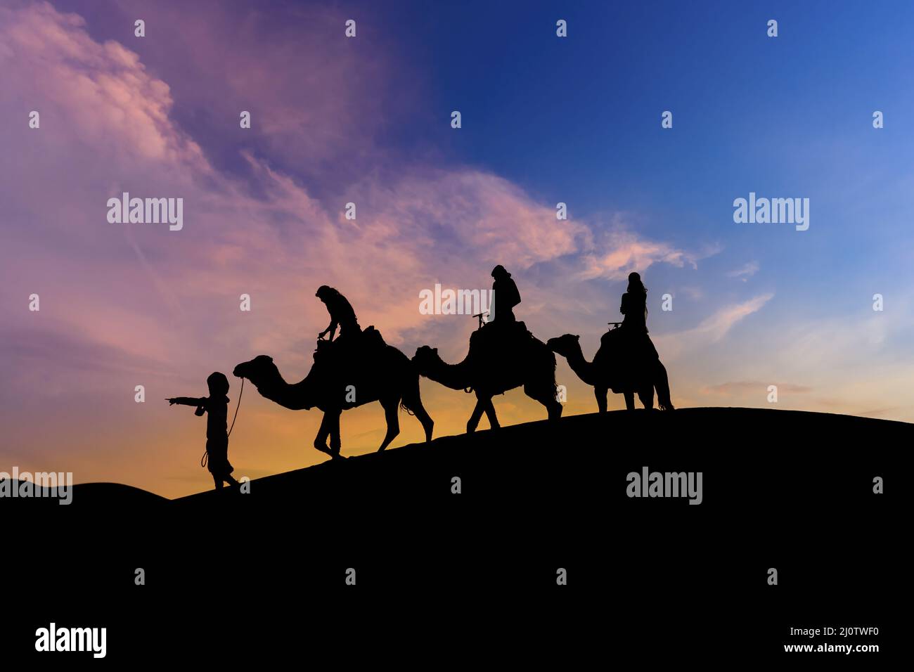 Three Riders And Their Handler Travel Through The Saharan Desert On Their Camels In Morocco Stock Photo