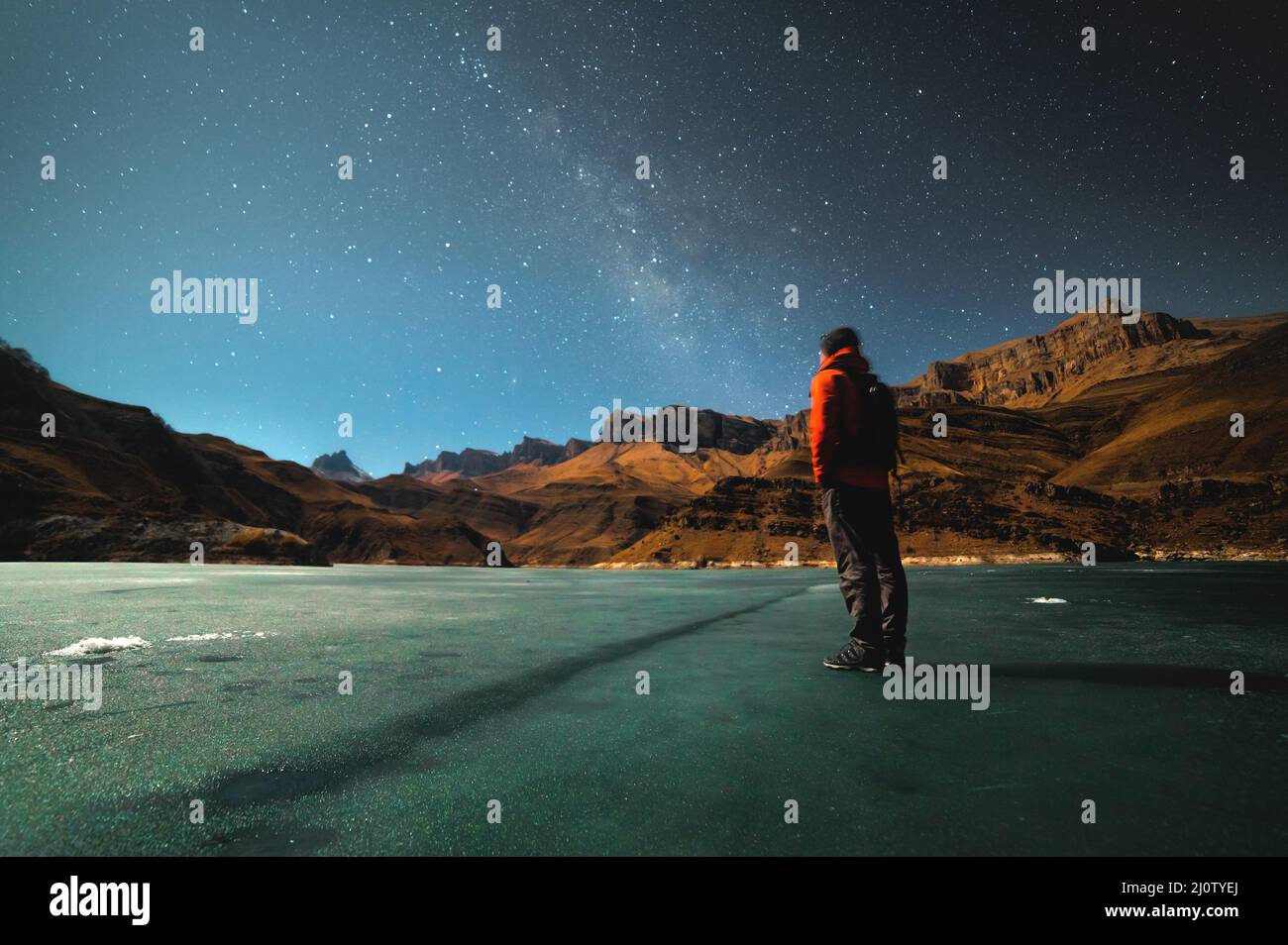 A brooding man stands on the surface of a frozen lake illuminated by moonlight high in the mountains against the backdrop of epi Stock Photo