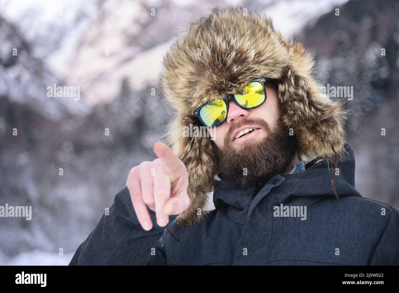 An attractive bearded man in a fur hat and sunglasses points his finger in the winter mountains in the woods and smiles Stock Photo