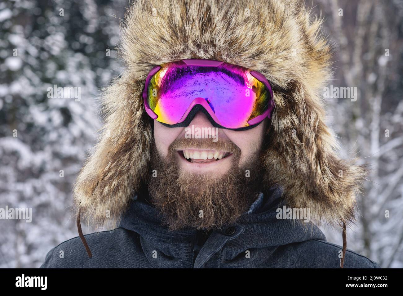 Close-up portrait of a bearded happy snowboarder skier in a ski mask with goggles and a fur big old-school hat on a background o Stock Photo