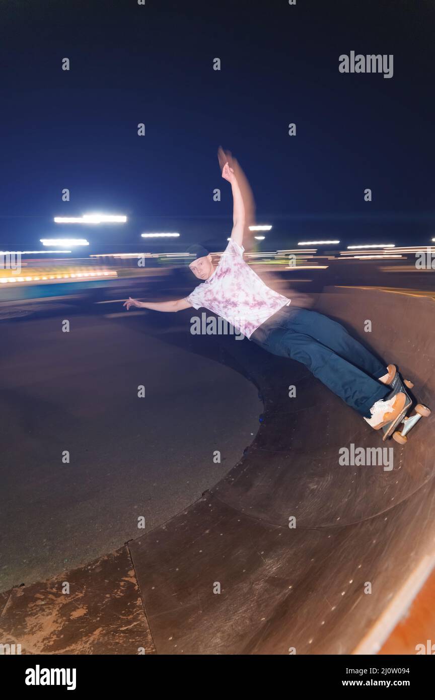 A young skateboarder rides at speed sideways along the ramp by spreading his hands to the sides. Night shot with long exposure a Stock Photo