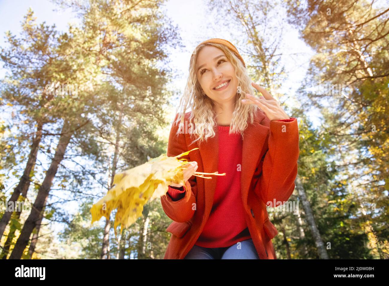 Attractive white Caucasian girl in red polto and orange hat is confused with a bouquet of leaves in her hands against the backgr Stock Photo