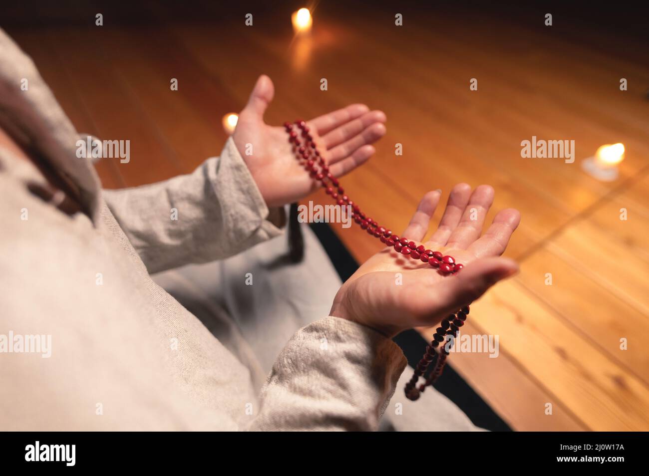 A close-up of the monk's hands hold red rosaries against the background of the wooden floor of the practice room and lit candles Stock Photo