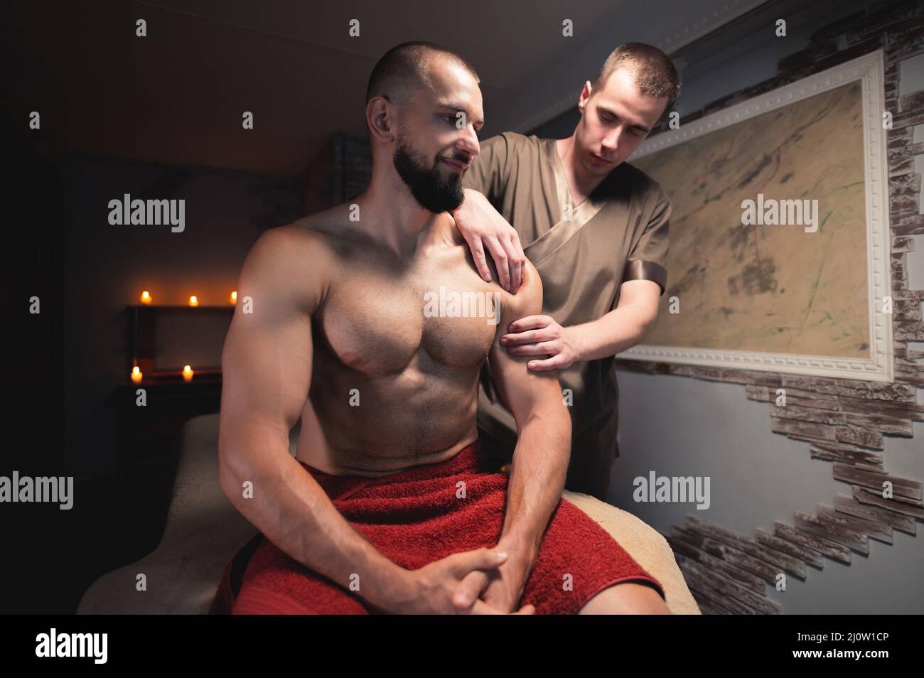 Male masseur does a sports shoulder massage to a muscular male athlete in a room with a contrasting dark light. Professional spo Stock Photo