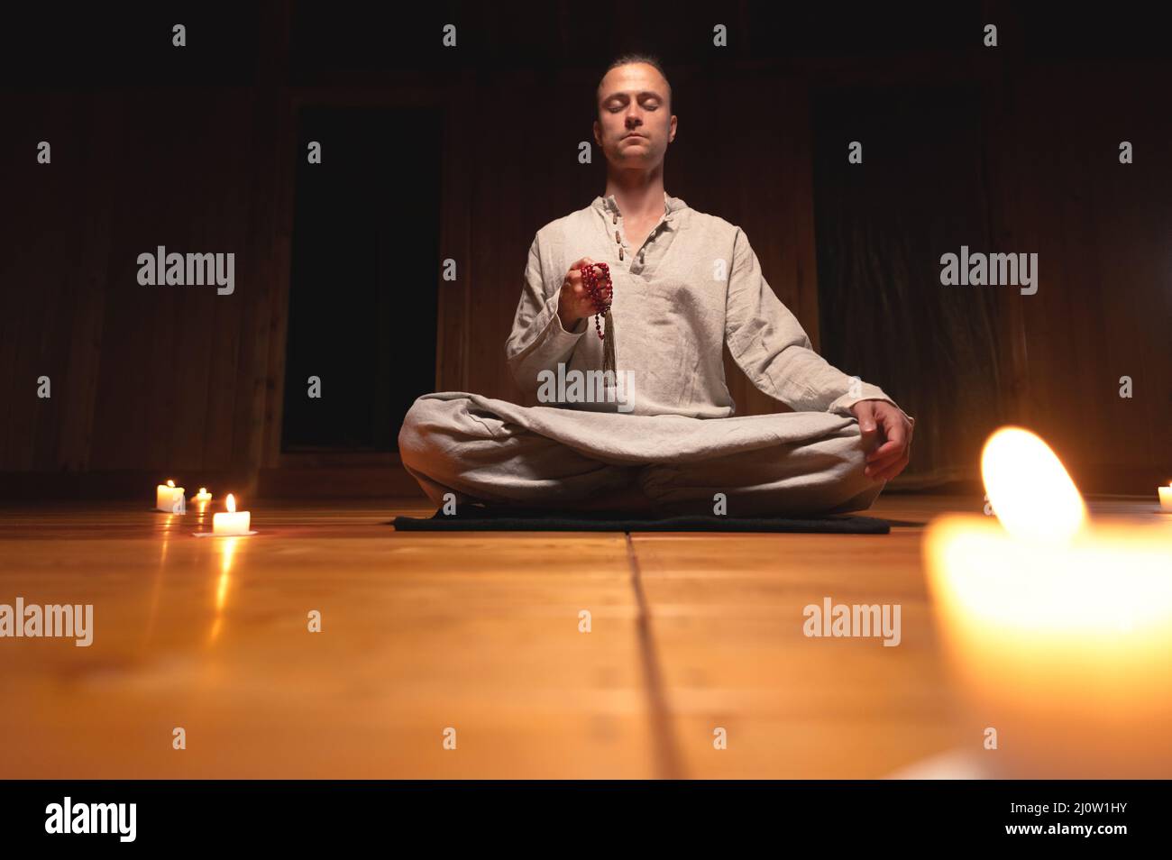 A man in clothes for practice and meditation sits in a lotus pose and holds red rosary to concentrate attention in a wooden room Stock Photo