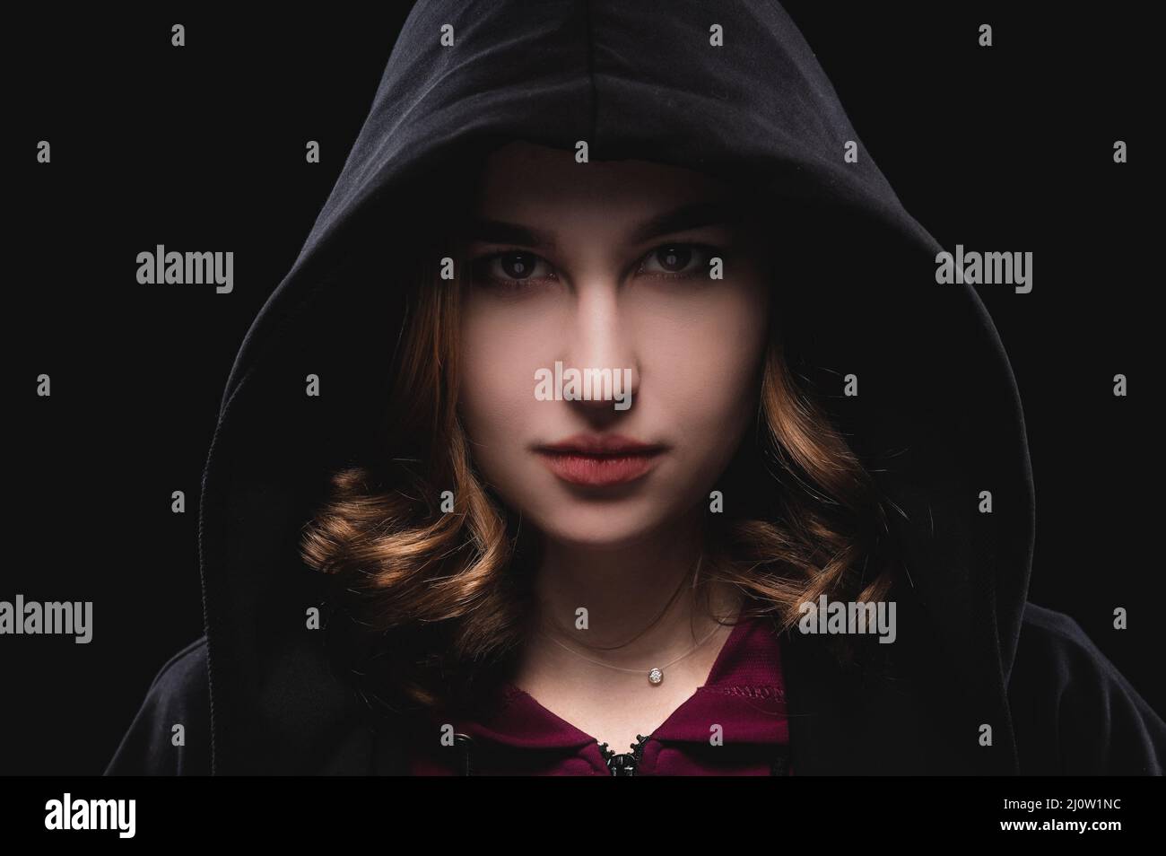 Close-up portrait of a secretive young girl in a deep dark hood on a black background. The concept of secrecy of secrets and peo Stock Photo