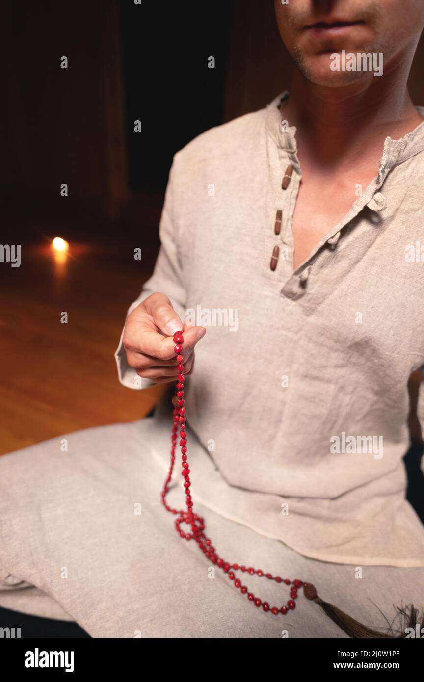 A man in clothes for practice and meditation sits in a lotus pose and holds red rosary to concentrate attention in a wooden room Stock Photo