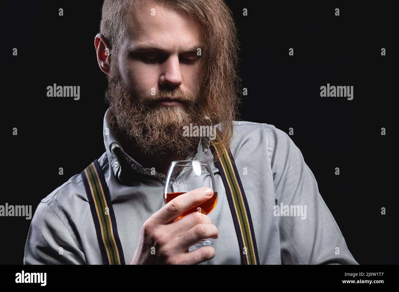 An attractive man with a long bang beard and a mustache sitting against the wall sniffs the scent of an alcoholic beverage in a Stock Photo