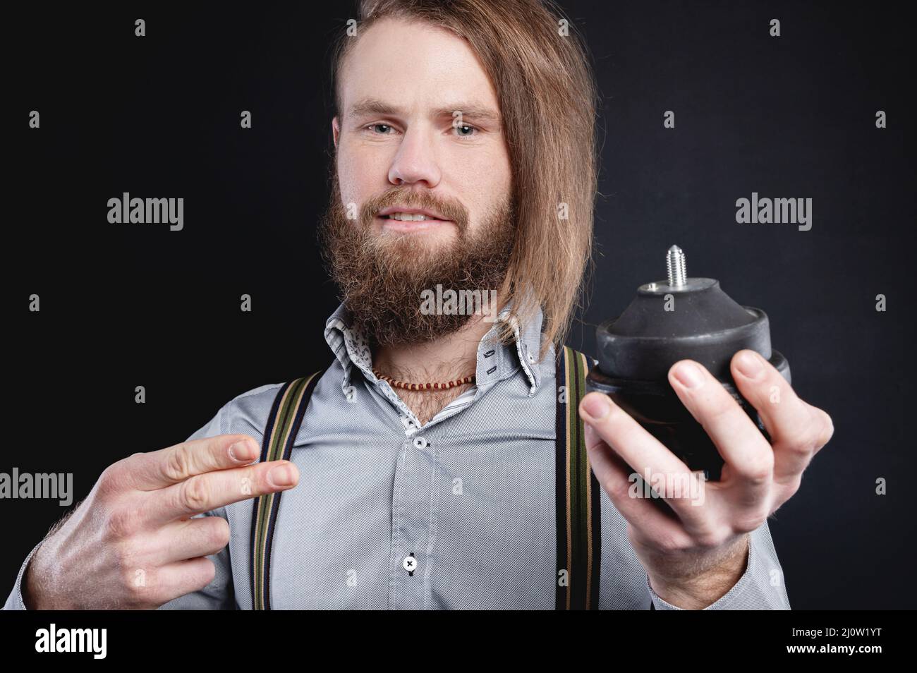 Portrait of a stylish bearded long-haired male salesman and dealer spare parts of the car in a shirt and suspenders. Holds engin Stock Photo