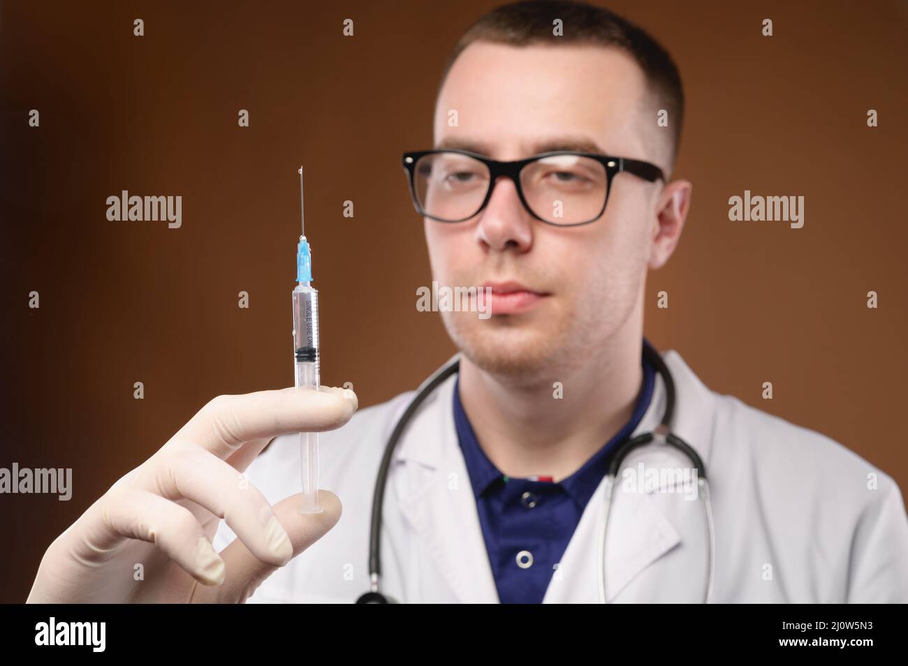 Young caucasian doctor man preparing to do a vaccination with a syringe. Studio shot on a brown background. Release air from the Stock Photo