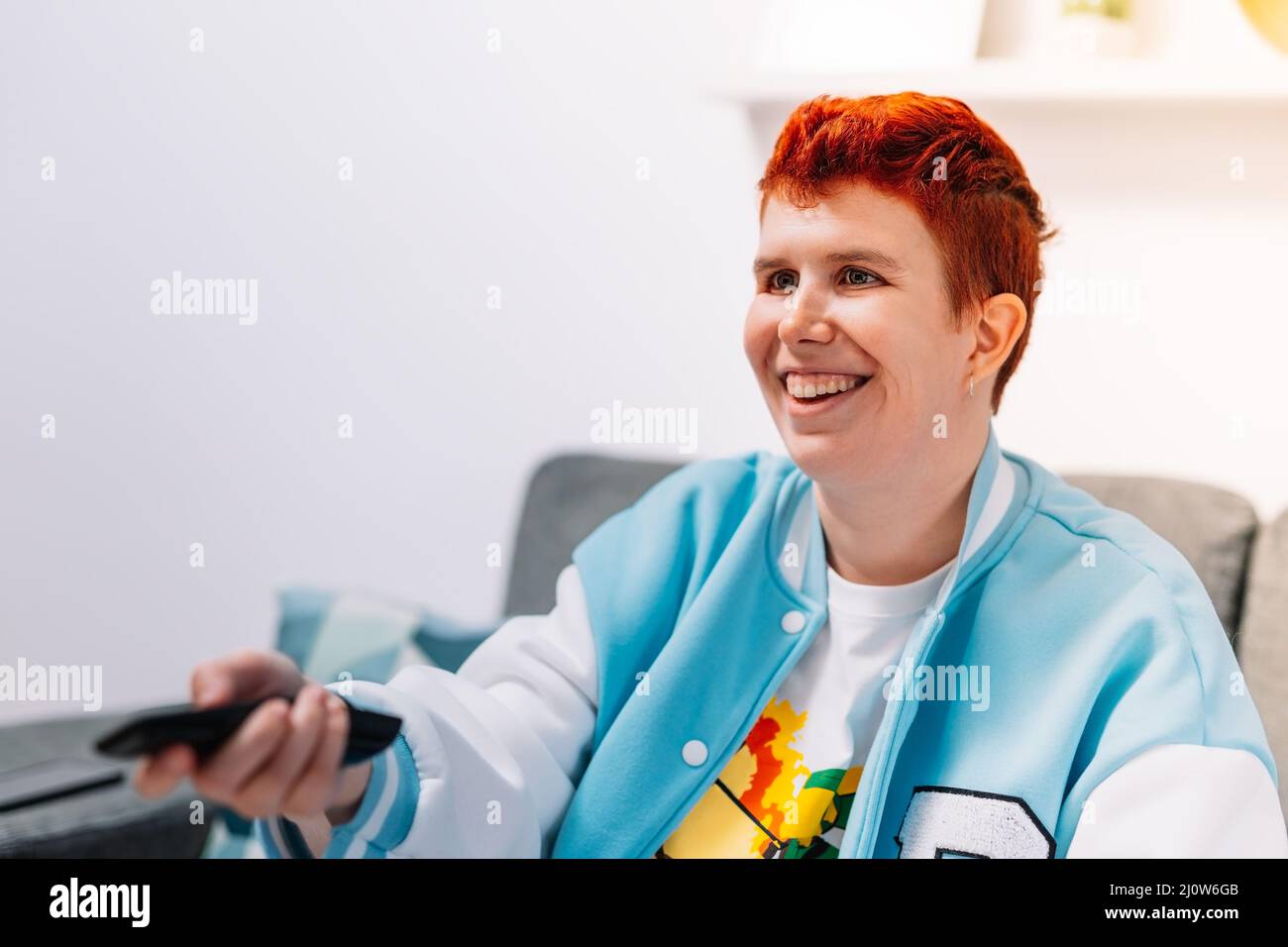 Close-up of a modern woman with short red hair, non-binary, changing the channel with the TV remote control. Stock Photo