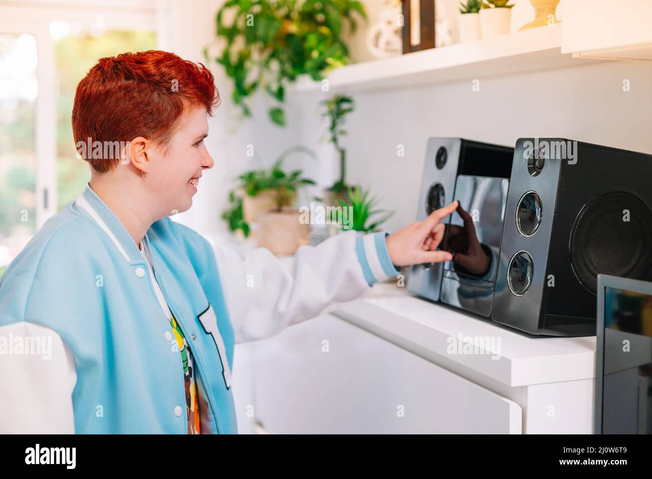 Modern, short-haired, red-haired, non-binary woman, turns on mini-system at home. Stock Photo
