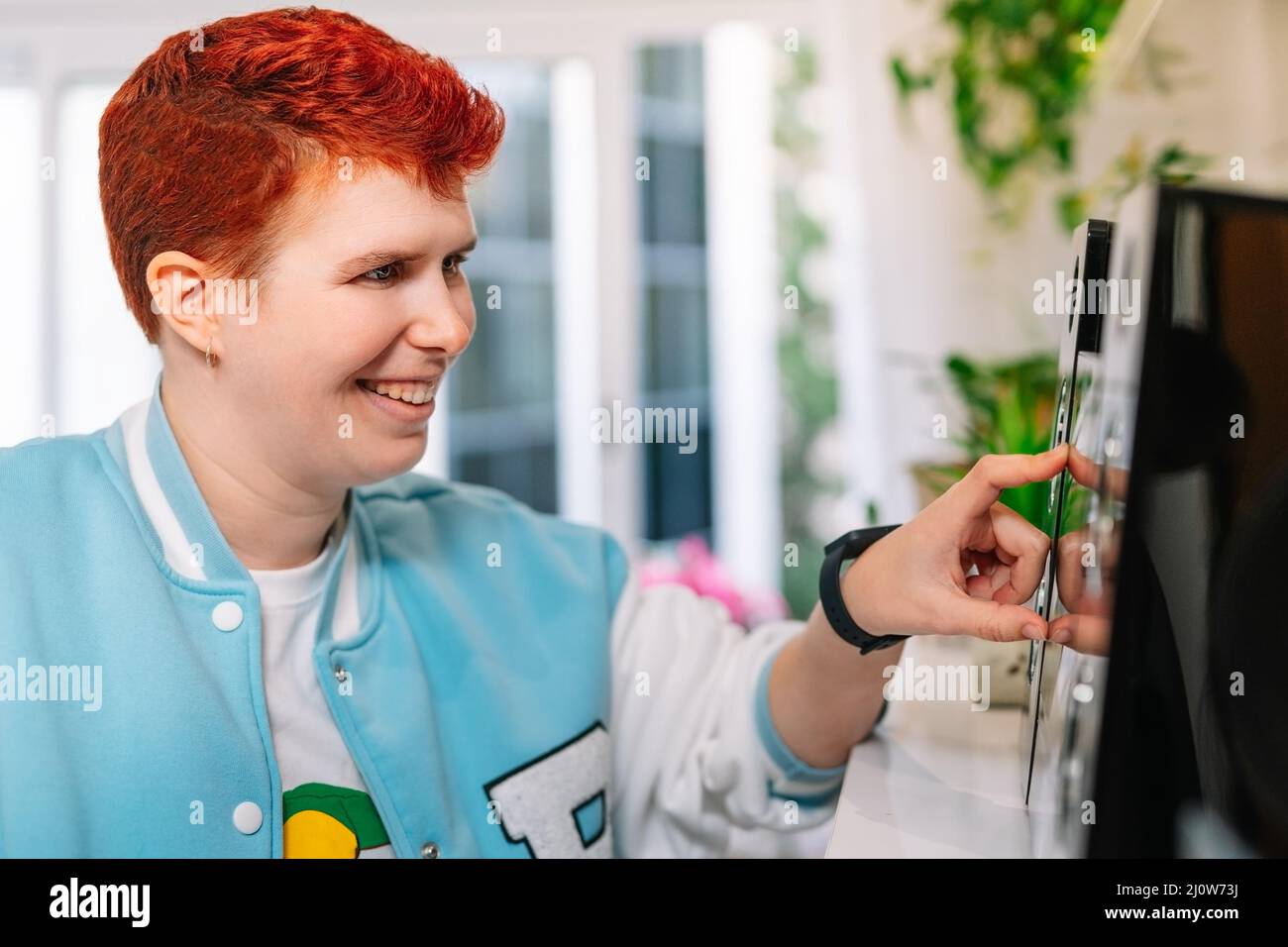 Close-up of a Modern, short-haired, red-haired, non-binary woman, turns on mini-system at home. Stock Photo