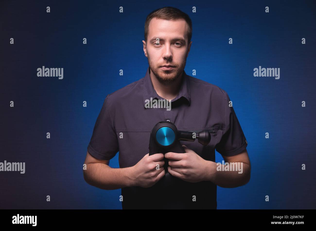 Portrait of a confident professional masseur with a percussion massager in his hands. Low key, blue background. Shock wave massa Stock Photo