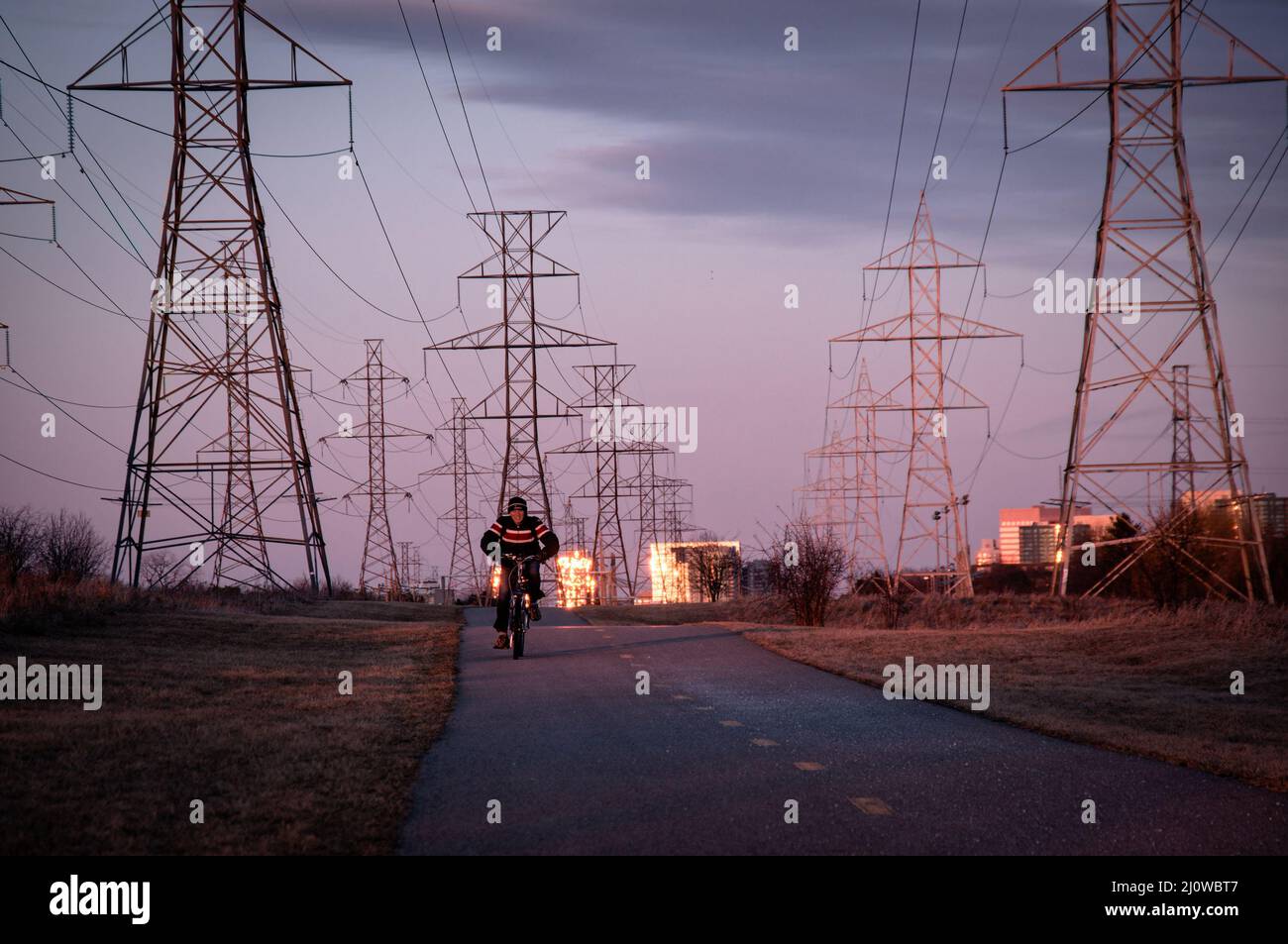 A man riding a bicycle along the walking trail beside the power line towers row in front of spectacular setting sun reflection in windows of high rise Stock Photo