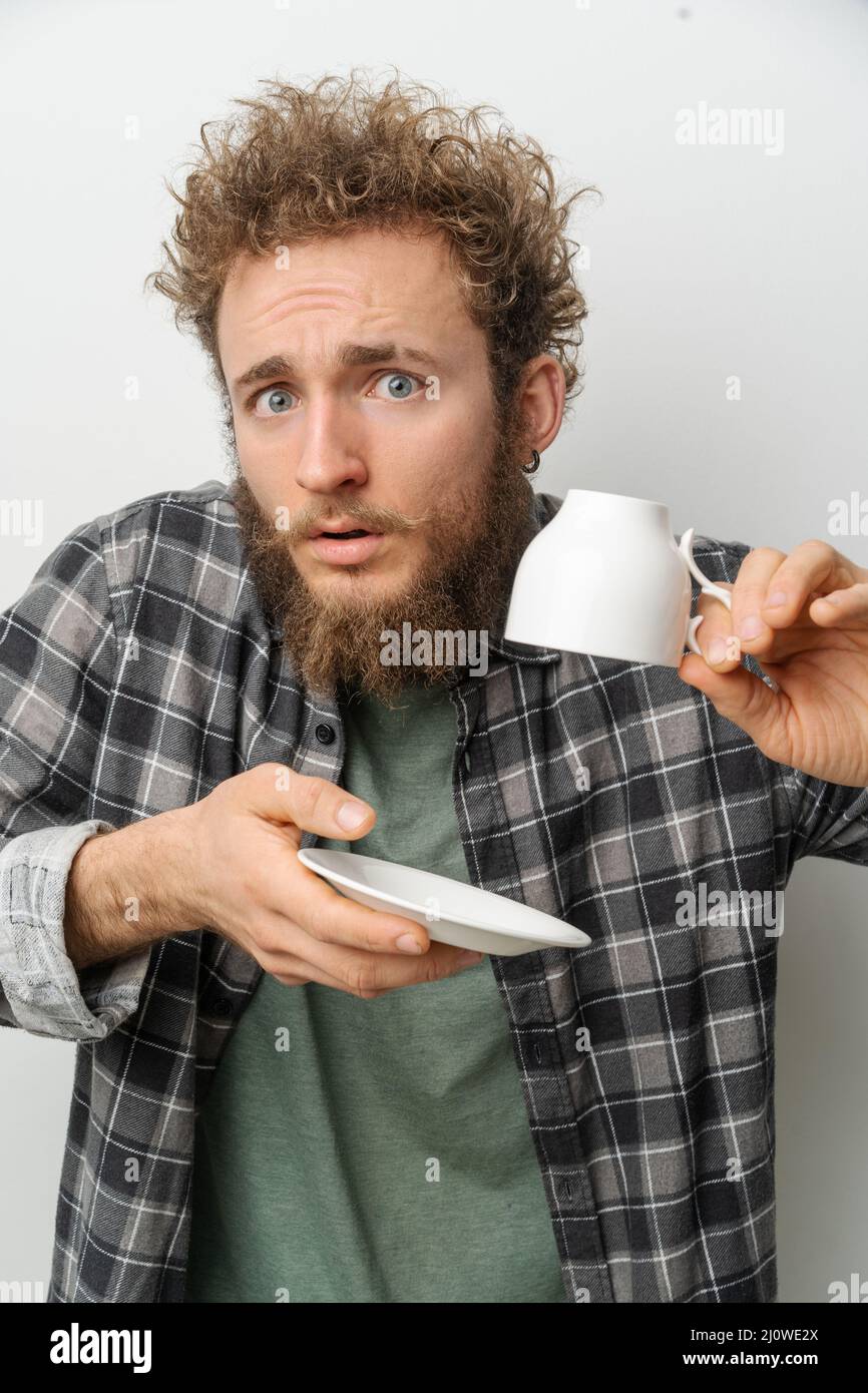 Surprised Man Holding White Empty Cup Upside Down and Saucer. Empty Coffee Cup. White Background. Close-up. Copy Space Stock Photo