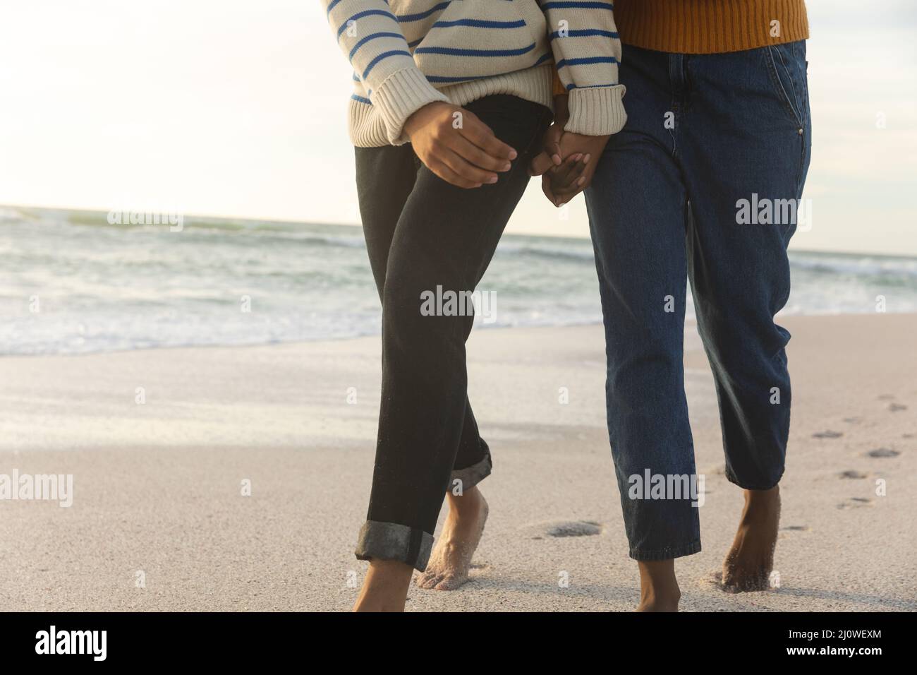 Low section of biracial couple holding hands while walking together on shore at beach during sunset Stock Photo