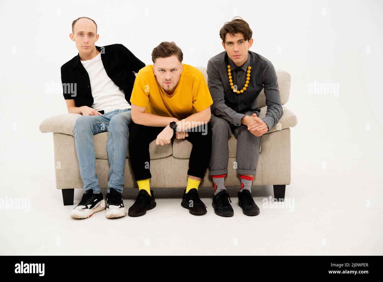 Young Guys Sit on a Sofa on a White Background and Look Together at the Camera. Three Friends are Sitting on the Couch and Watch Stock Photo