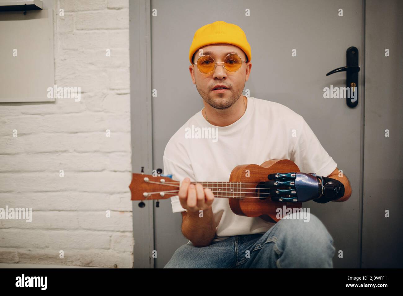 Young disabled man with artificial prosthetic hand plays on guitar at shop. Stock Photo