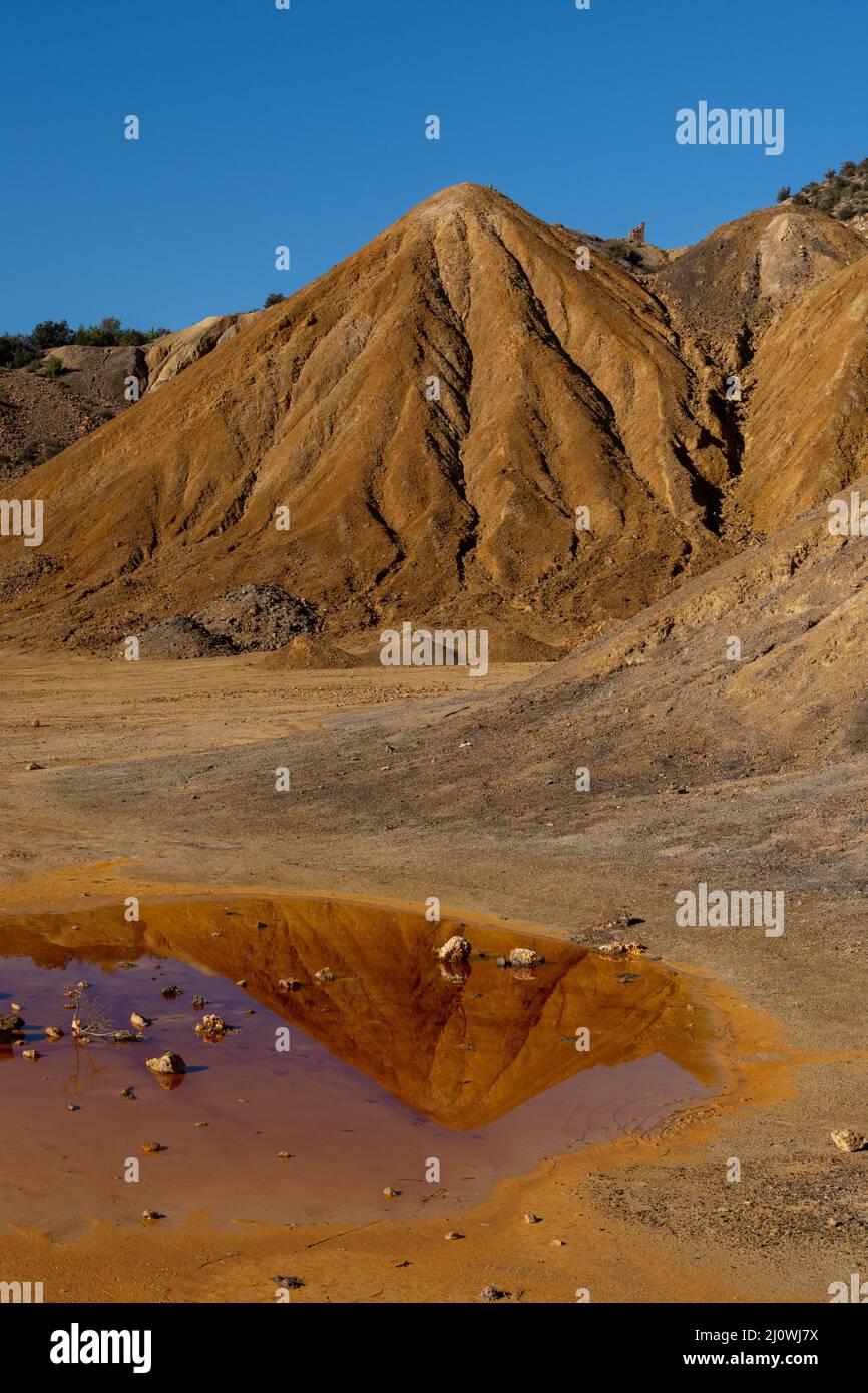 Abandoned mining grounds with eroded hillsides and reflections in a dirty pool of polluted water Stock Photo
