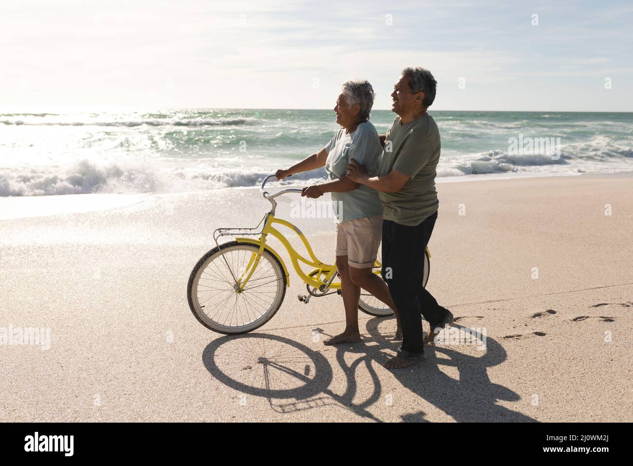 Side view of multiracial senior couple walking with bicycle on shore at beach during sunny day Stock Photo