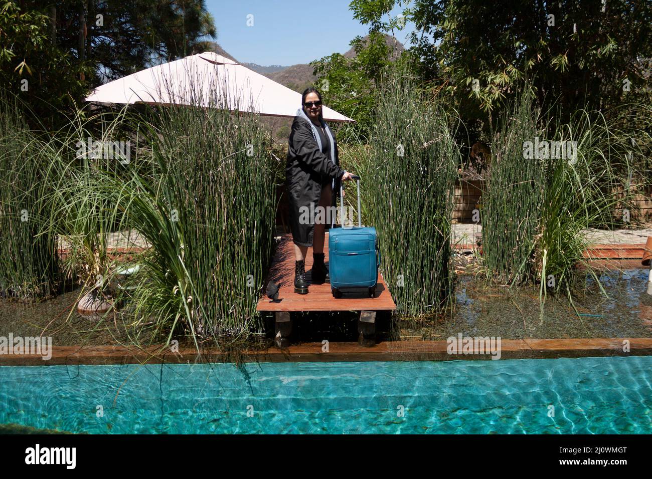 Caucasian Mexican woman dressed in a bathing suit, cape, boots and sunglasses arriving at a natural outdoor pool directing a wheeled suitcase over a w Stock Photo